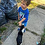 child, boy, toddler, pet_cat, black_and_white_cat, cat, sidewalk, concrete, trash_bags, garbage, grass, outdoor, daylight, blue_shirt, sneakers, smile, hand_petting, curious, curb, urban