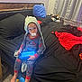 child, toddler, pacifier, tablet, pajamas, spiderman, hood, bed, blanket, pillow, curtain, nightstand, carpet, bare_feet, toy, bedroom, red_blanket, black_sheet, blue_pillow, clutter