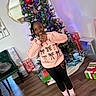 child, girl, christmas_tree, holiday, presents, indoors, smiling, heart_hands, sweatshirt, leggings, slippers, socks, wooden_floor, mirror, chair, decorations, lights, festive, happy, home