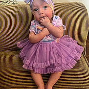 Jayla is registered to the contest to win money with this photo: baby, barefoot, child, clothing, couch, cute, feet, girl, hands, happy, headband, indoor, infant, person, portrait, purple_dress, sitting, smiling, striped_couch, toddler