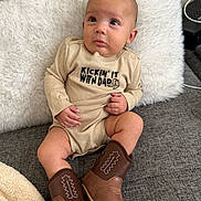 Zaire is registered to the contest to win money with this photo: baby, infant, onesie, cowboy_boots, couch, pillow, indoor, child, cute, portrait, sitting, clothing, footwear, furniture, young_child, expression, looking_up, skin, head, hand
