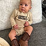 baby, infant, onesie, cowboy_boots, couch, pillow, indoor, child, cute, portrait, sitting, clothing, footwear, furniture, young_child, expression, looking_up, skin, head, hand