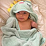 Zaire is registered to the contest to win money with this photo: baby, child, infant, towel, hood, dinosaur, yellow_spikes, blanket, bed, smile, face, cute, soft_texture, wrapped, skin, person, lying_down, indoors, cozy, happy