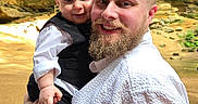 Zeke is registered to the contest to win money with this photo: man, baby, child, holding, smile, beard, white_shirt, black_vest, outdoor, rocky_background, happy, portrait, person, family, nature, sunlight, cute, closeup, head, face