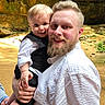 man, baby, child, holding, smile, beard, white_shirt, black_vest, outdoor, rocky_background, happy, portrait, person, family, nature, sunlight, cute, closeup, head, face