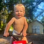 child, toddler, outdoor, toy_vehicle, honda, sunlight, blonde_hair, blue_eyes, smiling, bare_chest, diaper, handlebars, play, summer, daytime, backyard, trees, shed, fun, young_child