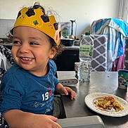 Zakaria a rejoint le concours — aidez-le/la à gagner de superbes lots ! toddler, child, crown, high_chair, smiling, food, plate, kitchen, table, water_bottle, cup, curly_hair, blue_shirt, indoor, happy, messy_face, seat, furniture, person, meal