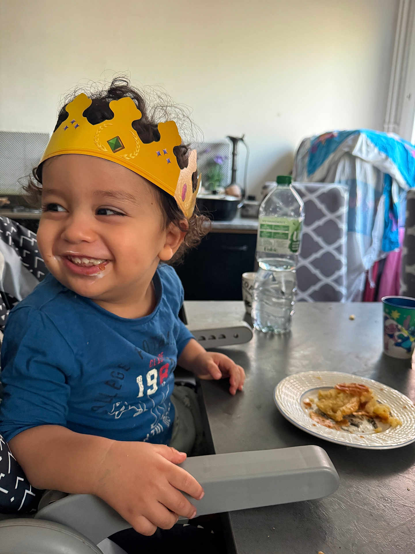 Zakaria a rejoint le concours — aidez-le/la à gagner de superbes lots ! toddler, child, crown, high_chair, smiling, food, plate, kitchen, table, water_bottle, cup, curly_hair, blue_shirt, indoor, happy, messy_face, seat, furniture, person, meal