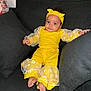 baby, child, yellow_outfit, headband, blue_eyes, couch, gray_cushions, daisy_pattern, sitting, barefoot, curly_hair, indoor, portrait, cute, fashion, comfortable, relaxed, smiling, person, soft_lighting