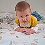 animal_print, baby, bed, blanket, blue_pants, child, cute, face, hands, home, indoor, infant, lying_down, person, playful, smiling, soft_toy, tongue_out, white_clothing, yellow_clothing