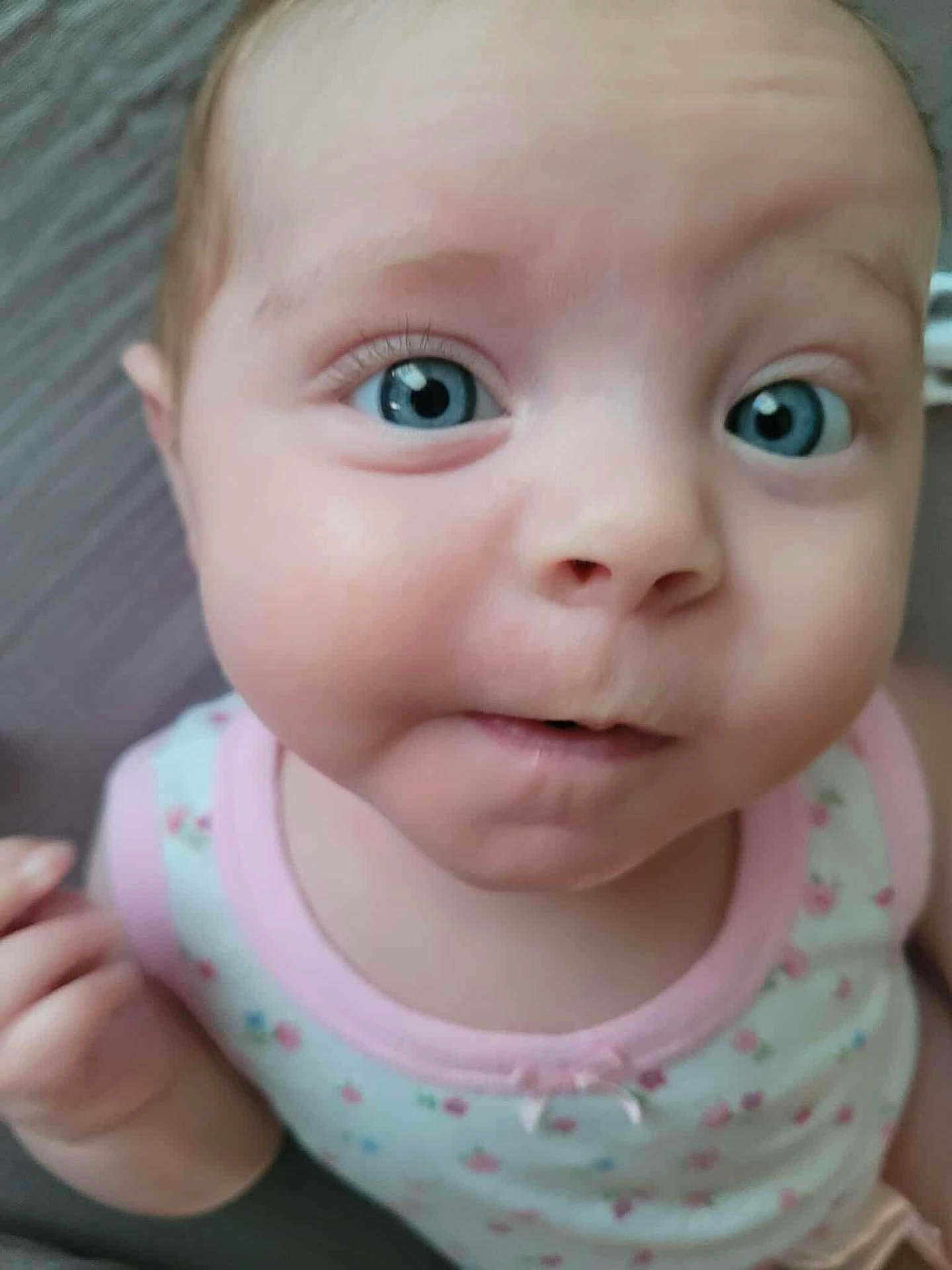 Jensyn is registered to the contest to win money with this photo: baby, close_up, face, blue_eyes, curious, infant, child, pink_clothing, floral_pattern, skin, portrait, cute, expression, indoor, person, young_child, head, small_hand, looking_up, soft_background