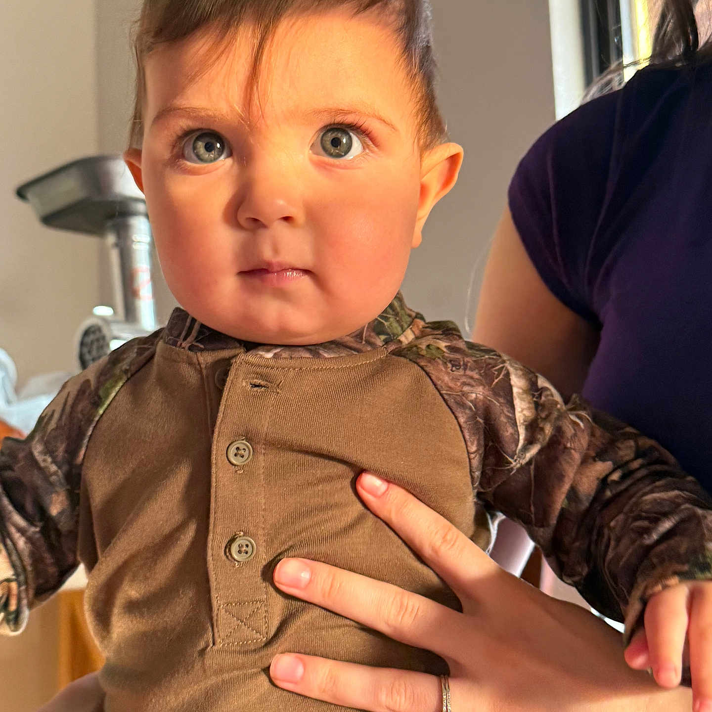 Alessio is registered to the contest to win money with this photo: adult_hand, baby, blue_eyes, buttoned_shirt, camouflage, child, clothing, expression, face, hand, holding, indoor, natural_light, person, portrait, ring, serious_face, short_hair, skin, young_child