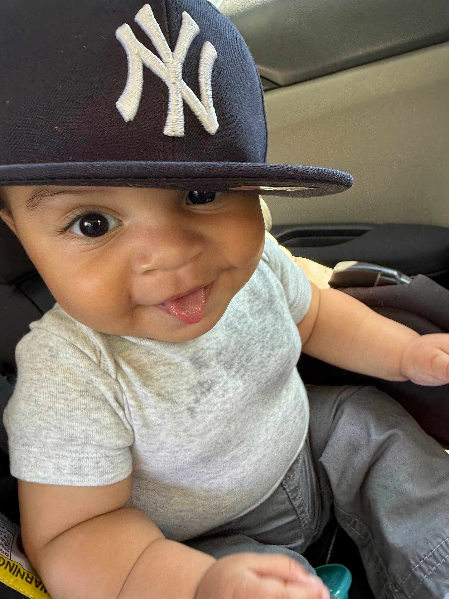 Julian joined the competition — help win amazing prizes! baby, child, face, tongue, cap, hat, ny_logo, gray_shirt, pants, car_seat, smiling, cute, playful, infant, portrait, closeup, indoor, person, young, casual_clothing