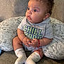 baby, child, infant, sitting, couch, pillow, animal_pattern, curly_hair, socks, shirt, bracelet, red_string, expression, wide_eyes, indoors, cozy, hands, clothing, cute, portrait
