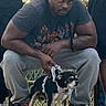 man, dog, outdoor, chain_link_fence, sneakers, crouching, harness, pet, casual_clothing, short_sleeves, grey_pants, black_headwear, muscular, person, animal, small_dog, daylight, photography, urban, people