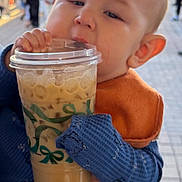 Ashton is registered to the contest to win money with this photo: baby, child, iced_coffee, drink, cup, hands, nails, street, outdoor, people, blurred_background, blue_sweater, orange_bib, face, portrait, daylight, urban, candid, cute, holding