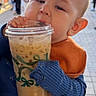 baby, child, iced_coffee, drink, cup, hands, nails, street, outdoor, people, blurred_background, blue_sweater, orange_bib, face, portrait, daylight, urban, candid, cute, holding