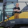child, boy, smiling, yellow_vehicle, tractor, agriculture, large_tire, outdoor, machinery, black_rail, clothing, short_sleeve_shirt, pants, blonde_hair, standing, daylight, farm_equipment, vehicle, happy, metal