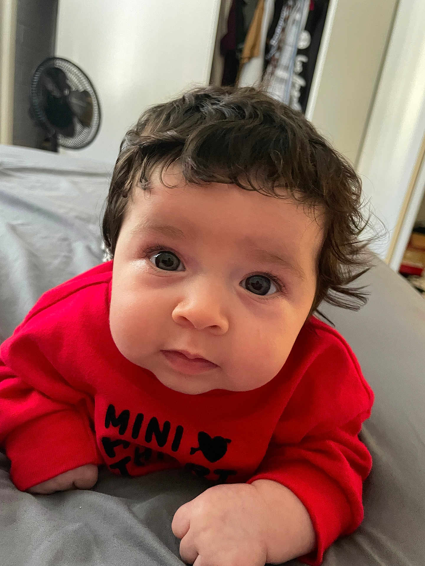 Aurora is registered to the contest to win money with this photo: baby, child, curly_hair, red_sweater, face, eyes, bed, blanket, hand, indoor, clothing, person, infant, cute, portrait, young_child, home, furniture, fan, expression