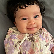 Aurora is registered to the contest to win money with this photo: baby, infant, smile, curly_hair, floral_outfit, pink, purple, blanket, cute, portrait, face, child, happy, lying_down, soft_texture, cozy, indoors, young_child, sleepwear, baby_clothes