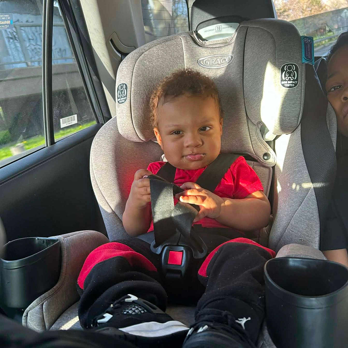 Messiah is registered to the contest to win money with this photo: toddler, child, car_seat, car_interior, seatbelt, red_shirt, black_pants, sneakers, window, sunlight, car, baby, person, seat, safety, cute, vehicle, travel, daylight, portrait