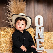 Cris is registered to the contest to win money with this photo: toddler, child, cowboy_hat, black_clothing, hay_bale, balloons, birthday, one_year, portrait, rustic, decor, indoor, cute, sitting, smiling, curly_hair, floor, wooden_background, celebration, boots