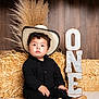 toddler, child, cowboy_hat, black_clothing, hay_bale, balloons, birthday, one_year, portrait, rustic, decor, indoor, cute, sitting, smiling, curly_hair, floor, wooden_background, celebration, boots