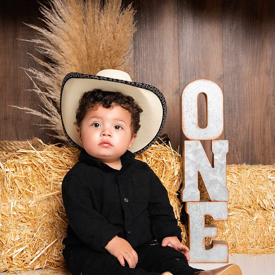 Cris is registered to the contest to win money with this photo: balloons, birthday, black_clothing, boots, celebration, child, cowboy_hat, curly_hair, cute, decor, floor, hay_bale, indoor, one_year, portrait, rustic, sitting, smiling, toddler, wooden_background