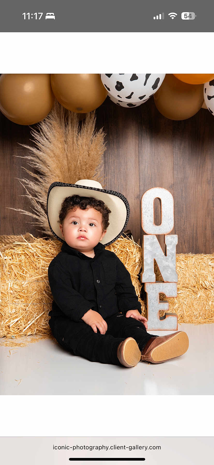 Cris is registered to the contest to win money with this photo: toddler, child, cowboy_hat, black_clothing, hay_bale, balloons, birthday, one_year, portrait, rustic, decor, indoor, cute, sitting, smiling, curly_hair, floor, wooden_background, celebration, boots