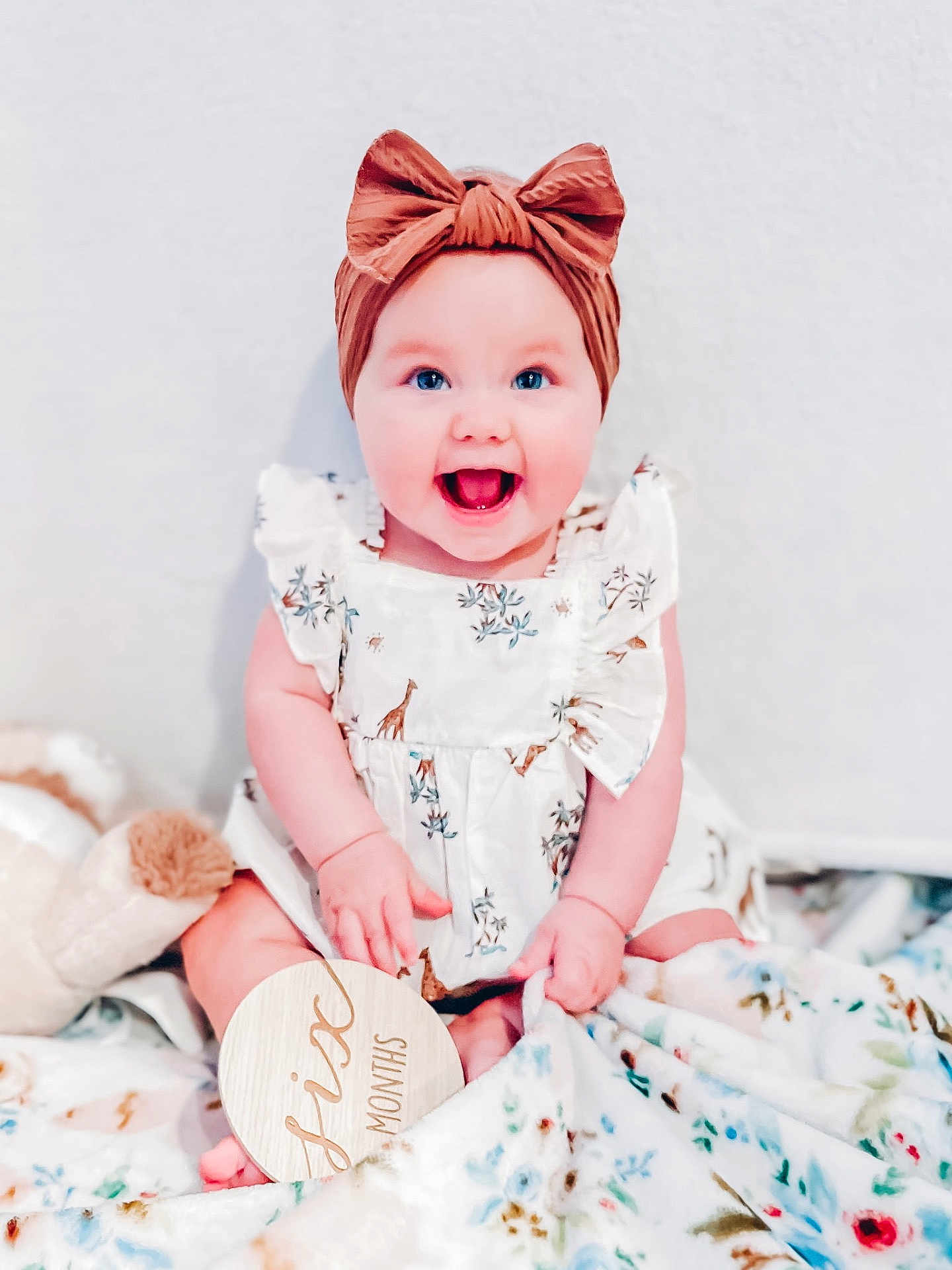 Londyn is registered to the contest to win money with this photo: baby, child, smiling, headband, bow, dress, giraffe_print, blue_eyes, blanket, floral, wooden_sign, sitting, cute, happy, indoor, infant, portrait, hands, feet, soft_toys