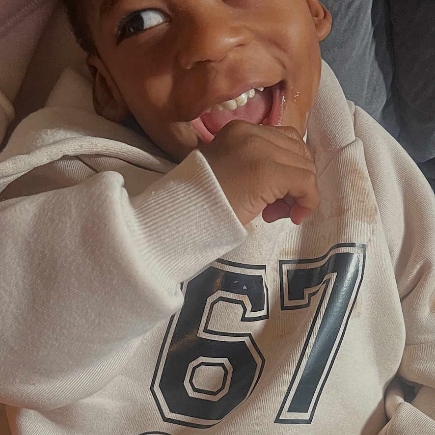 Champ is registered to the contest to win money with this photo: bedding, candid, casual, child, closeup, clothing, couch, cozy, expressive_eyes, hand_in_mouth, hoodie, indoor, kid, laugh, number_67, pillow, playful, portrait, smiling, teeth