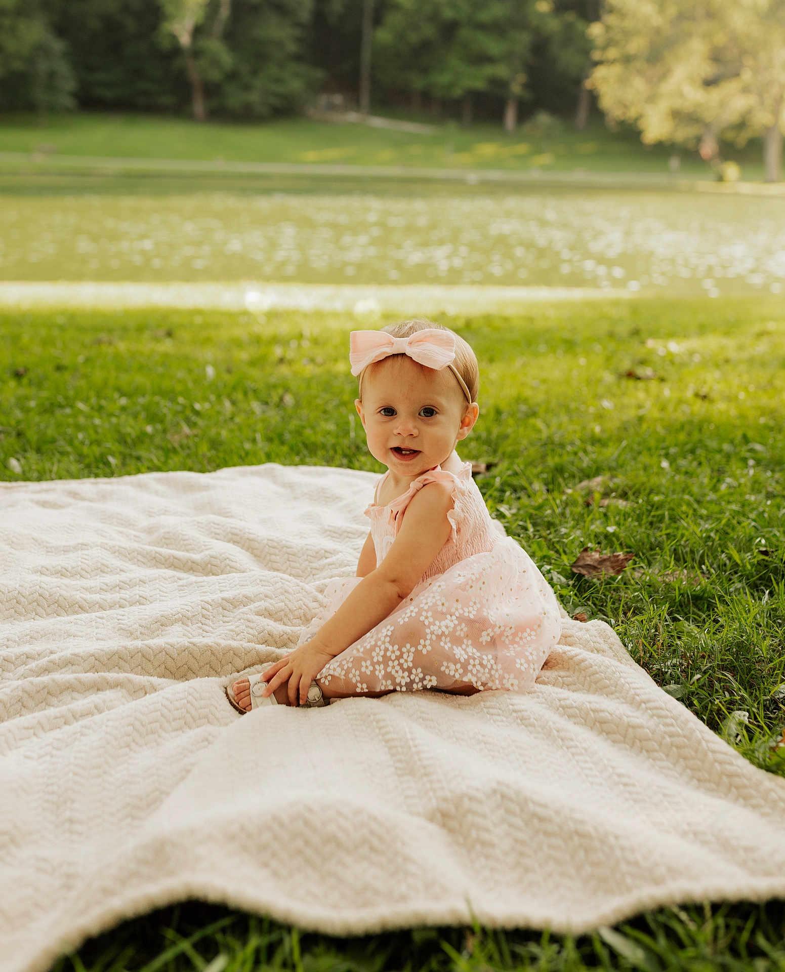Kora joined the competition — help win amazing prizes! baby, child, girl, pink_dress, bow, blanket, grass, lake, outdoor, nature, smile, portrait, sitting, cute, sunlight, greenery, young_child, headband, happy, summer