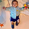 Sebastian is registered to the contest to win money with this photo: baby, child, smiling, play_mat, blue_jacket, cat_patch, shorts, booties, toy, colorful, indoor, happy, face, person, infant, lying_down, cute, childrens_toy, patterned_mat, cozy