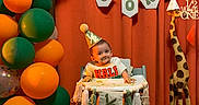 Sebastian joined the competition — help win amazing prizes! baby, child, high_chair, party_hat, balloons, orange_curtain, banner, celebration, giraffe_statue, half_birthday, smiling, festive, decorations, indoor, person, toddler, wooden_giraffe, colorful, happy, event