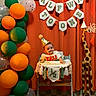 baby, child, high_chair, party_hat, balloons, orange_curtain, banner, celebration, giraffe_statue, half_birthday, smiling, festive, decorations, indoor, person, toddler, wooden_giraffe, colorful, happy, event