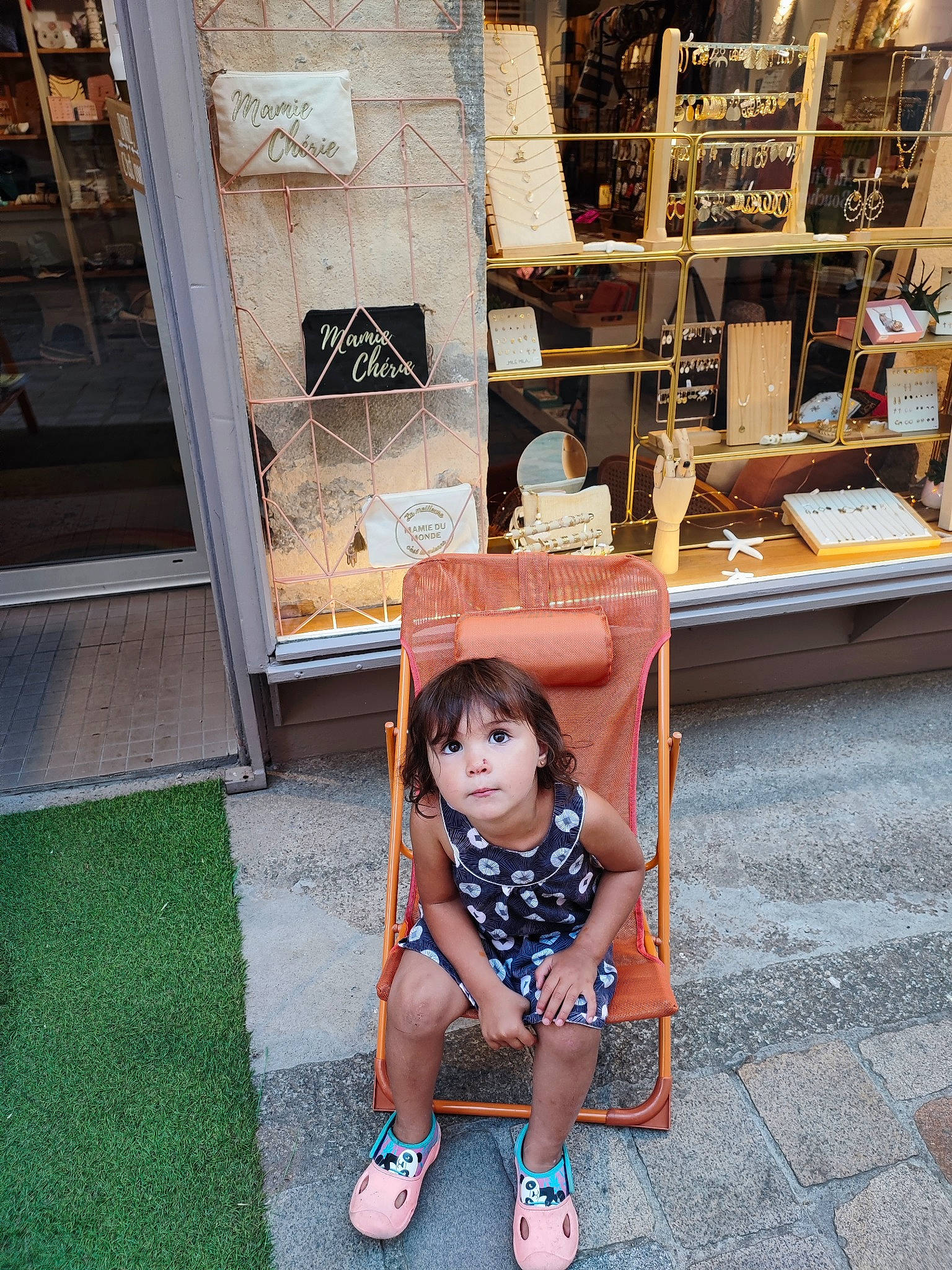 Anna participe au concours pour gagner de l'argent avec cette photo : chair, city, dress, eyewear, flooring, human_leg, leisure, person, plant, sandal, shelf, shelving, shoe, sidewalk, sitting, t_shirt, table, thigh, toddler, tree