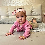 Ema is registered to the contest to win money with this photo: baby, child, infant, crawling, bed, pink_clothing, headband, indoor, person, young_child, cute, face, eyes, hands, legs, blanket, pillow, furniture, wall, expression