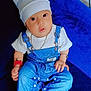accessories, adorable, baby, blue_background, bracelet, chain, child, cute, denim_overalls, hat, indoor, infant, looking, pacifier, papers, plush_surface, sitting, socks, table, white_tshirt