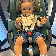 Oussaïd a rejoint le concours — aidez-le/la à gagner de superbes lots ! baby, blue_sneakers, car_seat, child, clothing, curious_expression, curly_hair, denim_shorts, floor_tiles, footwear, indoor, pacifier, person, safety, seatbelt, sitting, straps, toddler, toy, young_child