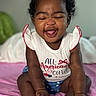 Jaliyah is registered to the contest to win money with this photo: baby, child, smiling, sitting, curly_hair, red_bows, white_shirt, text_on_clothing, blue_shorts, sparkly_shoes, pink_blanket, indoors, cute, happy, portrait, infant, person, clothing, expression, hands