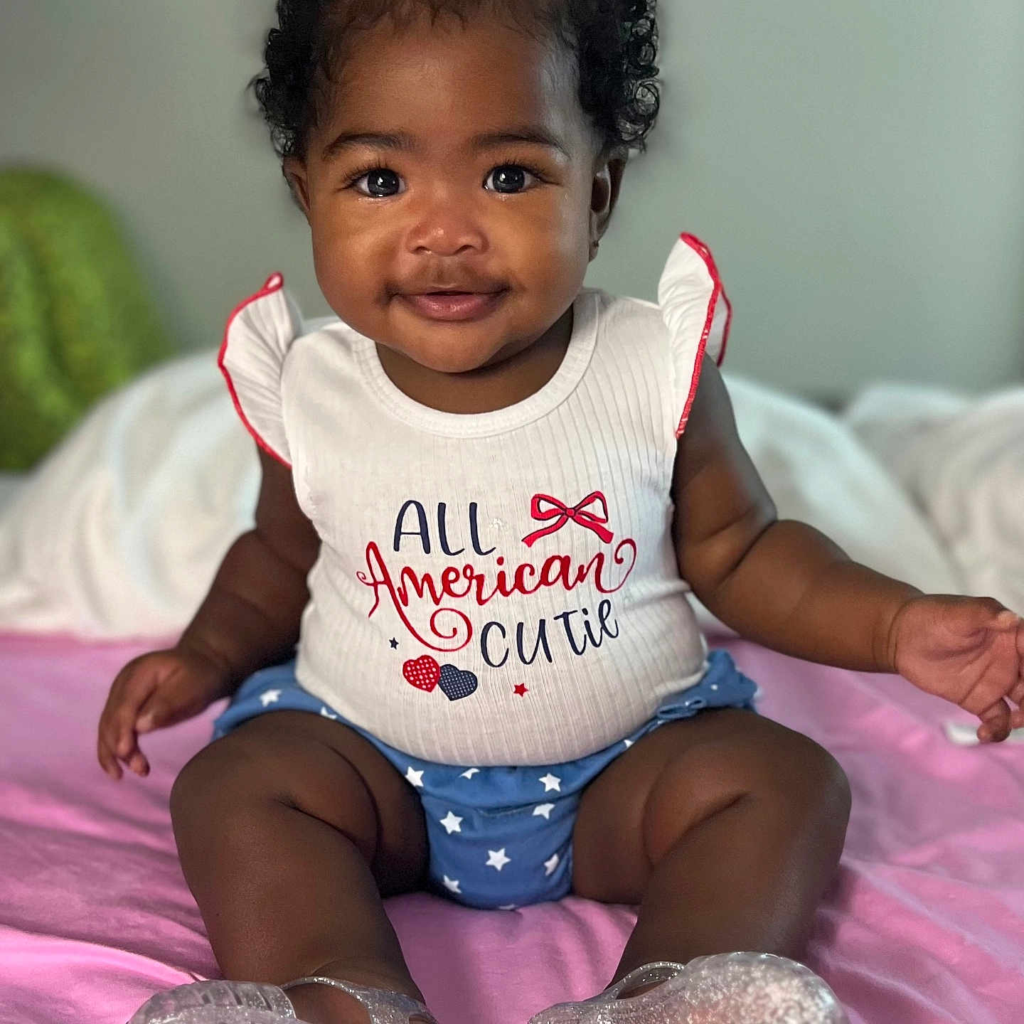 Jaliyah is registered to the contest to win money with this photo: baby, child, infant, cute, smiling, sitting, curly_hair, red_bows, white_top, blue_shorts, stars, pink_blanket, indoors, portrait, happy, young_child, footwear, transparent_shoes, person, apparel