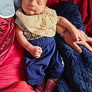 Thomas is registered to the contest to win money with this photo: adult_hand, baby, bib, blanket, blue_fabric, blue_pants, closeup, clothing, cozy, cute, finger, hand, indoors, infant, newborn, pink_socks, red_fabric, resting, sleeping, soft