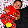 child, toddler, smiling, pajamas, red_clothing, yellow_socks, curly_hair, bed, blanket, patterned_background, lip_prints, happy, face, person, indoor, cute, playful, lying_down, colorful, cozy