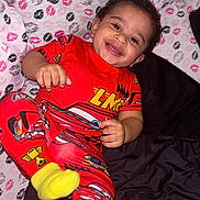 Kross is registered to the contest to win money with this photo: toddler, child, smiling, pajamas, red_clothing, race_car, bed, blanket, kissing_print, happy, face, curly_hair, yellow_socks, indoor, person, cute, playful, lying_down, cozy, bright_colors