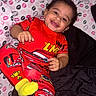 toddler, child, smiling, pajamas, red_clothing, race_car, bed, blanket, kissing_print, happy, face, curly_hair, yellow_socks, indoor, person, cute, playful, lying_down, cozy, bright_colors