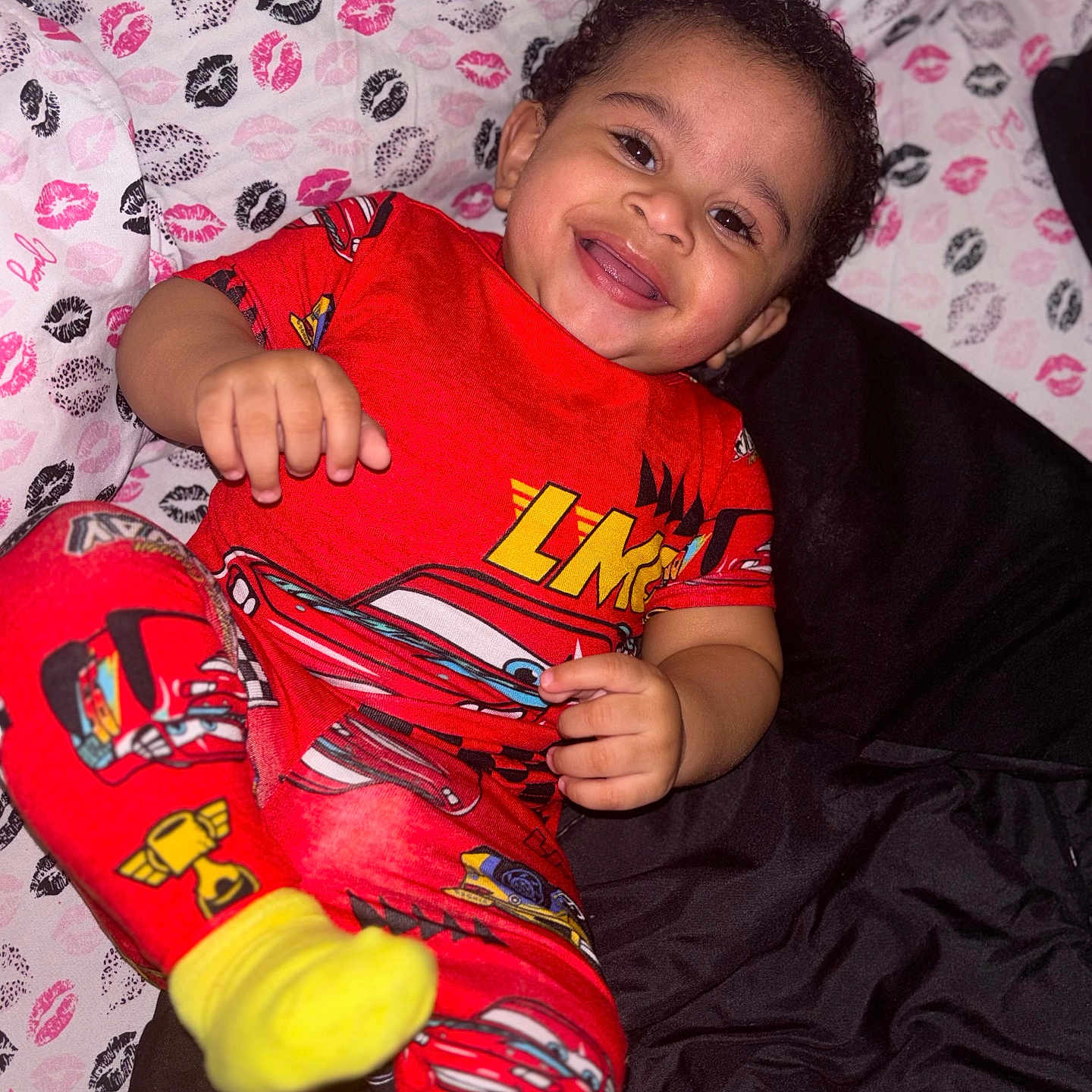 Kross is registered to the contest to win money with this photo: toddler, child, smiling, pajamas, red_clothing, race_car, bed, blanket, kissing_print, happy, face, curly_hair, yellow_socks, indoor, person, cute, playful, lying_down, cozy, bright_colors