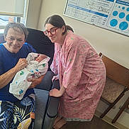 Elena joined the competition — help win amazing prizes! newborn, baby, woman, elderly_person, hospital_room, hospital_gown, blanket, chair, medical_board, window, glasses, floor, person, seated, smiling, holding, family, indoor, footwearless, portrait