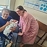 newborn, baby, woman, elderly_person, hospital_room, hospital_gown, blanket, chair, medical_board, window, glasses, floor, person, seated, smiling, holding, family, indoor, footwearless, portrait