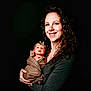 Astraea is registered to the contest to win money with this photo: woman, baby, infant, portrait, smile, curly_hair, headband, swaddle, holding, dark_background, studio_photo, happy, mother, child, person, clothing, jeans, long_sleeve, skin, face