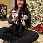 Zero is registered to the contest to win money with this photo: woman, dog, pomeranian, puppy, indoor, christmas_tree, holiday_lights, carpet, sitting, smiling, black_dog, white_dog, pet, home, decor, casual_clothing, leggings, shoes, wooden_sign, dog_bed